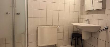 Un baño con cabina de ducha de vidrio, un lavamanos blanco, un taburete pequeño y un radiador.