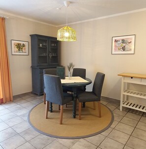 A dining area with a round table, four chairs, a decorative hanging light, a cabinet, and two framed artworks on the walls.