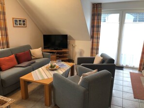 A cozy living room with a sofa, armchair, and a coffee table. A flat-screen TV is mounted on the wall, and there is a window with curtains.