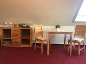 A room with a wooden desk, two chairs, a bookshelf, a small table with a plant, and a window with natural light.