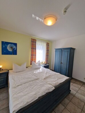 A bedroom with a bed, a nightstand, a lamp, a wardrobe, and a window with curtains. - Darßer Weg 36 Whg. 9 - Holiday apartment near the beach (Zingst)
