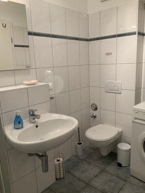 A modern bathroom with a white sink, toilet, and bidet. There is a mirror above the sink, a soap dispenser, and a toilet paper holder. The walls are tiled with a blue stripe, and there is a Siemens washing machine in the corner.