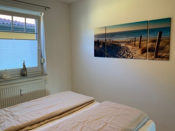 A bedroom with a bed, a radiator, a window with blinds, and a beach scene painting on the wall.