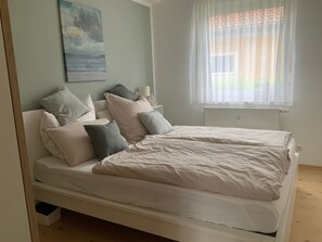 Un dormitorio con cama, almohadas, una mesita de noche, una lámpara y una ventana con cortinas.