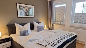 Uma cama bem arrumada com um edredom branco acolchoado, um travesseiro com um desenho náutico e um cobertor com a frase "HOME SWEET HOME" bordada.