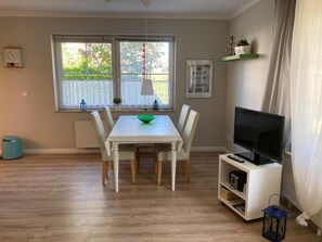 Un coin repas avec une table et des chaises blanches, un saladier vert et un téléviseur posé sur un meuble blanc.