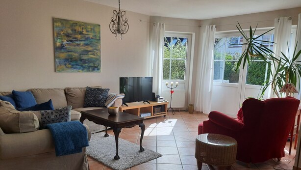 Ein Wohnzimmer mit einer Couch, einem roten Sessel, einem Couchtisch, einem Fernseher und einem großen Fenster mit Vorhängen.