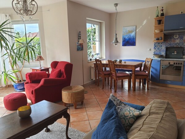 Un salon avec un fauteuil rouge, une table à manger avec des chaises, une cuisine avec des meubles bleus et une grande fenêtre.