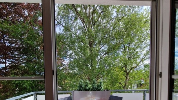 Un balcón con una mesa y sillas, una puerta de cristal y vistas a árboles.