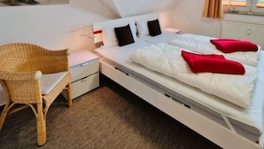 Un dormitorio con cama, una silla, una mesita de noche, una ventana y un cuadro colgado en la pared.