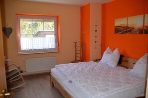 Ein Schlafzimmer mit einem großen Bett, einem Fenster mit Jalousien, einem Heizkörper, einem Holzstuhl und einem an der Wand befestigten Regal.