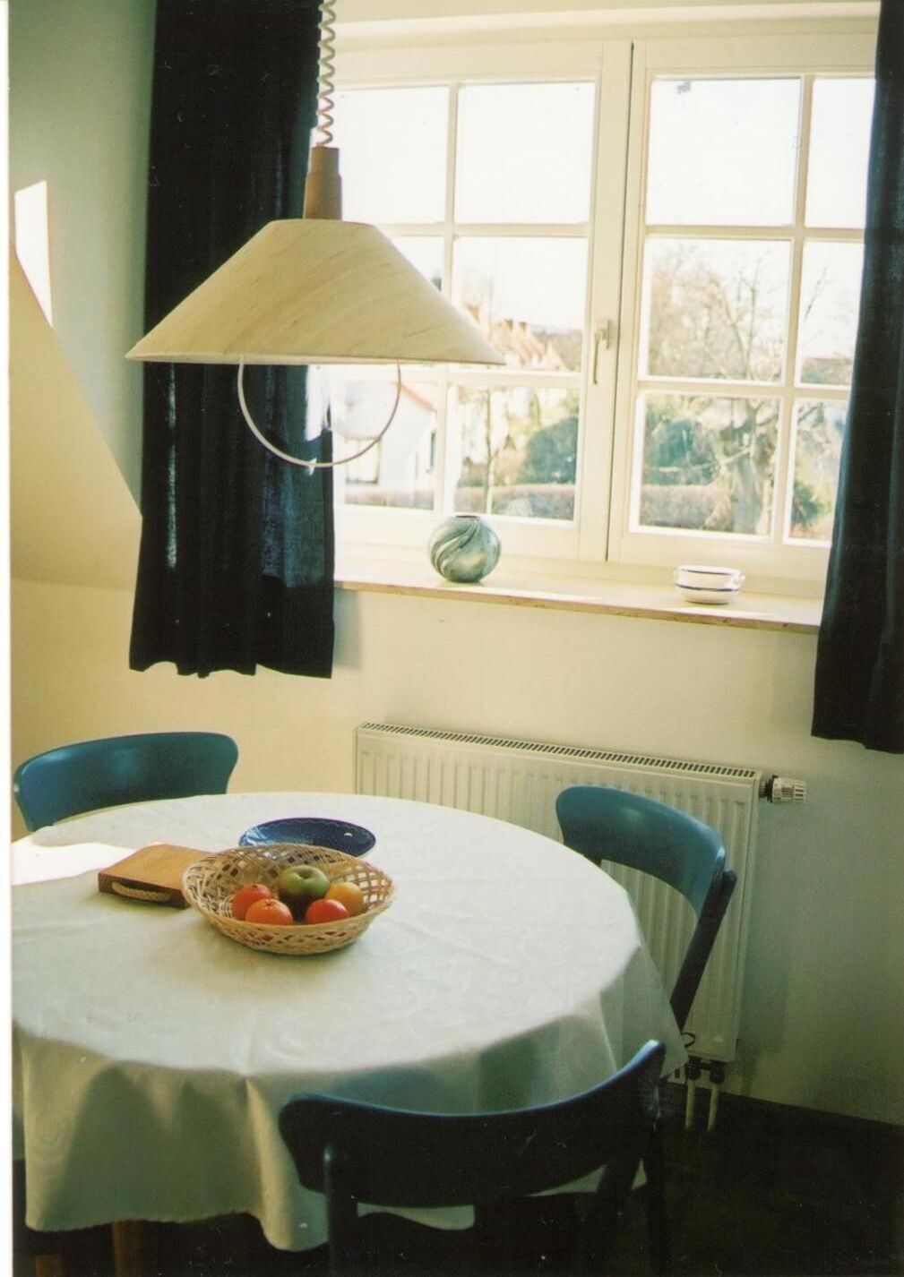 A dining area with a round table, chairs, a basket of fruit, a vase, and a window with curtains.