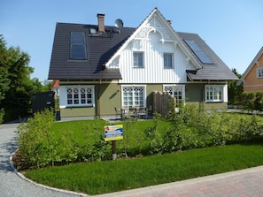 Ein zweistöckiges Haus mit geneigtem Dach, heller Fassade und einem kleinen Vorgarten mit Rasen und Sträuchern.