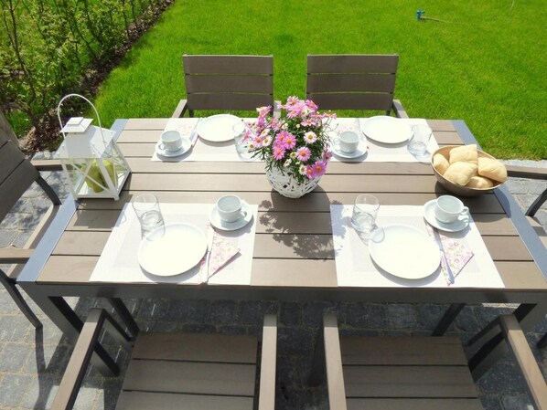 A wooden table set for two with white plates, cups, and a vase of pink flowers.