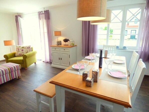 A dining area with a wooden table set for four, white chairs, and a window with curtains.