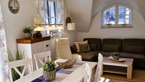 Un salon chaleureux avec une table à manger, un canapé et un petit coin cuisine agrémenté d’une horloge et de plantes.