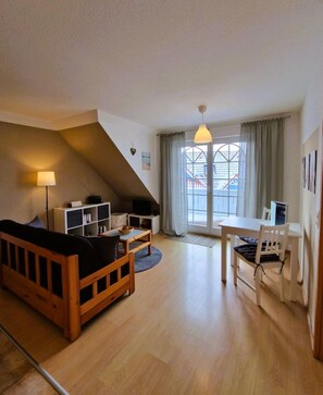 A cozy loft-style living room with a wooden sofa, a dining table with chairs, a bookshelf, and a large window with curtains.