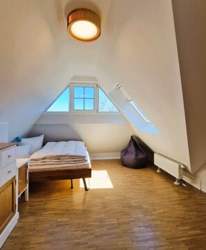 Ein Schlafzimmer mit schrägverdeckter Decke, einem Bett, einem Fenster, einem Heizkörper und einer Deckenleuchte.