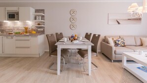 Una cucina moderna con pensili e basi bianche e un forno a microonde integrato. C'è un'area pranzo con un tavolo bianco e sedie marroni. La stanza presenta un divano beige e un tavolino da caffè bianco.