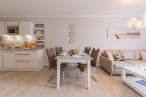 Eine moderne Küche mit weißen Schränken und einer eingebauten Mikrowelle. Es gibt einen Essbereich mit einem weißen Tisch und braunen Stühlen. Der Raum verfügt über ein beiges Sofa und einen weißen Couchtisch.