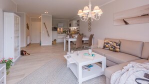 Un soggiorno moderno con un divano bianco, un tavolino da caffè e un lampadario. Si nota una zona pranzo con tavolo e sedie, e una cucina sullo sfondo.