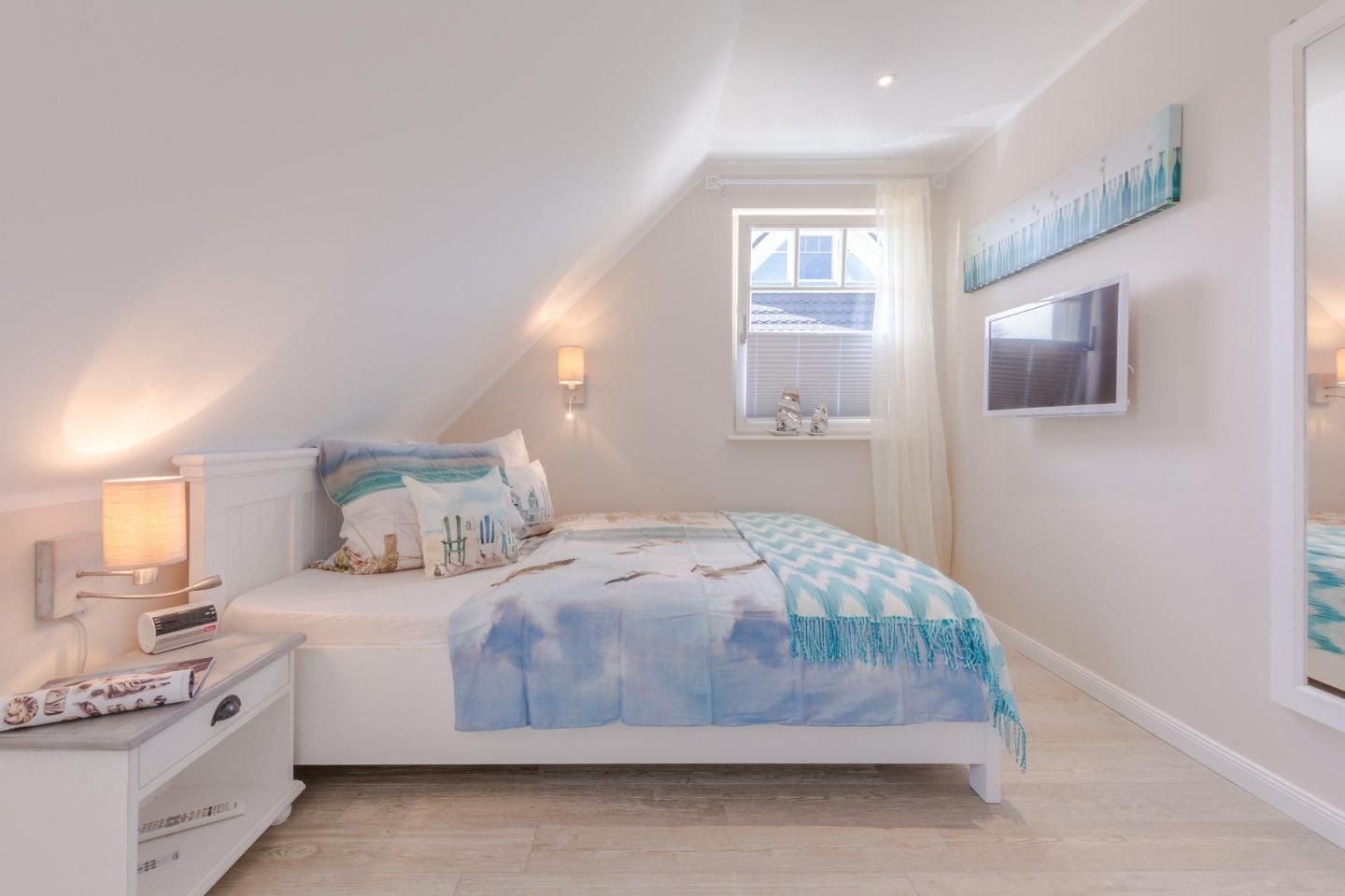 A bedroom with a sloped ceiling, a bed with a blue and white patterned bedspread, a bedside table with a lamp, and a window with blinds.