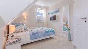 A bedroom with a sloped ceiling, a bed with a blue and white bedspread, a bedside lamp, a window with blinds, and a wall-mounted TV. - Birkmaase 1a "Abendklang" - comfortable semi-detached house "Abendklang" is quiet (Zingst)