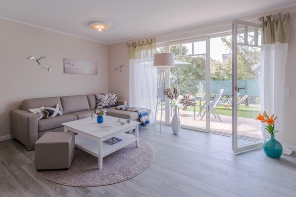 A modern living room with a sofa, coffee table, and a view of a garden through large sliding glass doors.