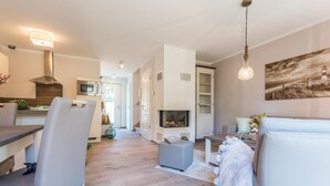 A modern living room with a fireplace, a dining area with a kitchen in the background, and a cozy seating area with a lamp.