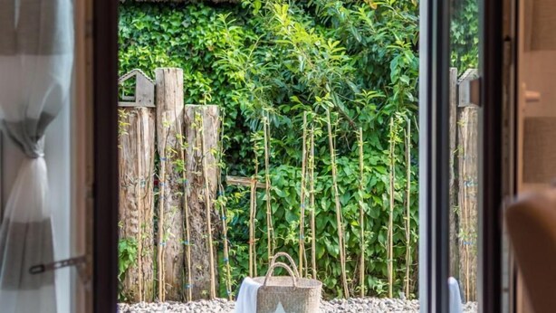 A view through a glass door to an outdoor area with a wooden deck, a thatched roof structure, and lush greenery.