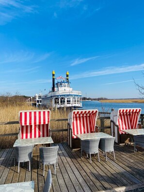 A deck with wicker chairs and tables, overlooking a body of water with a steamboat in the background. - Rämel 38 Whg. 15 "Strandkieker" - A place to dream and linger (Zingst)