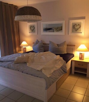 A neatly made bed with striped bedding, two bedside tables with lamps, and three framed pictures on the wall. - Rämel 38 Whg. 15 "Strandkieker" - A place to dream and linger (Zingst)