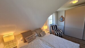 Ein ordentlich eingerichtetes Schlafzimmer mit einem Bett, Kissen, einem Sessel, einem Nachttisch mit Lampe und einer Wanduhr.