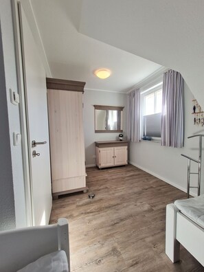 Una habitación con piso de madera, una puerta blanca, un ventanal con cortinas, un espejo y un armario.
