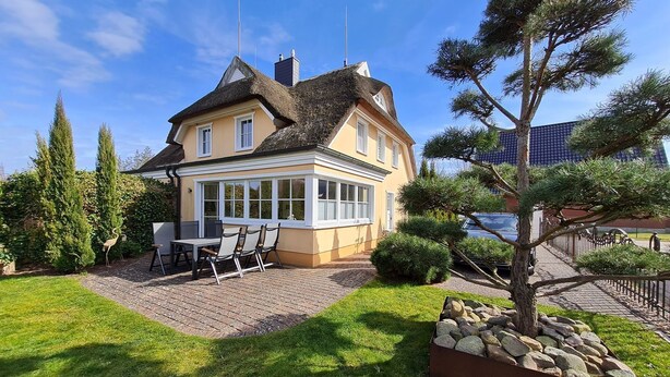 Ein zweigeschossiges Haus mit Wintergarten, einer Terrasse mit Tisch und Stühlen sowie einem gepflegten Garten.