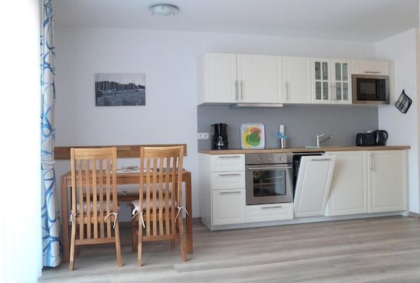 Une cuisine compacte avec des meubles blancs, un four à micro-ondes, un four traditionnel et un évier. On y trouve une table à manger en bois avec des chaises. Un tableau est accroché au mur.