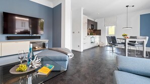 En moderne stue med et fladskærms-tv, en sofa, et sofabord med bøger og en skål med frugt, samt et spiseområde med bord og stole.