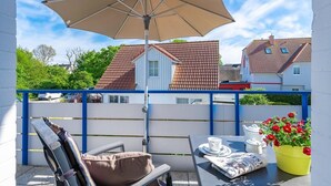 En balkon med et bord dækket til to, en stol, en stor parasol og en potteplante med røde blomster.