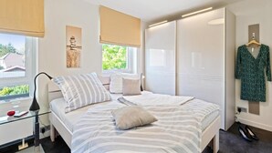 Et soveværelse med seng, puder, et natbord med lampe, et vindue med persienner, et klædeskab og et ophængt tøj.