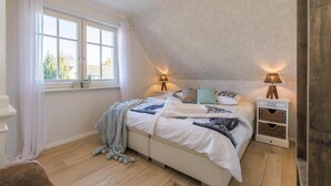 A bedroom with a bed, two bedside tables with lamps, a window with curtains, and a wooden floor.