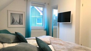 Ein Schlafzimmer mit einem Bett, Kissen, einem Fernseher, einem Fenster mit Vorhängen und einem gerahmten Bild an der Wand.