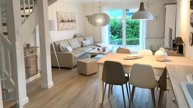 Ein modernes Wohnzimmer mit einer Couch, einem Esstisch und Stühlen. Eine Treppe befindet sich links, und ein großes Fenster bietet Blick auf viel Grün.