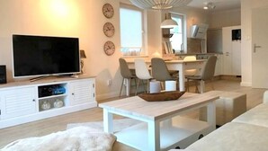 Ein modernes Wohnzimmer mit Flachbildfernseher, einem Essbereich mit Stühlen, einer Küche im Hintergrund und einer großen Pendelleuchte.