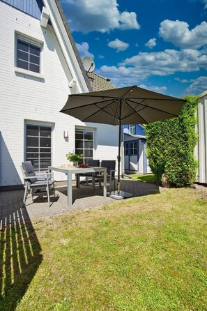 Eine Terrasse mit Tisch und Stühlen, einem großen Sonnenschirm sowie ein weißes Gebäude mit Fenstern und einer Sat-Anlage.