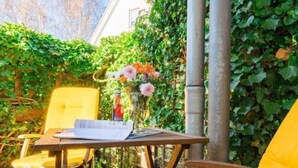 Een houten tafel met een menukaart en een fles wijn, omringd door gele stoelen in een tuin met muurranken.