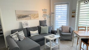Een moderne woonkamer met een grijze hoekbank, een kleine witte tafel en een eetgedeelte met een houten tafel en stoelen.