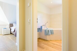 A modern bedroom with a bed, bedside table, and a small window. The bathroom features a bathtub, a shower, and a towel rack. - Kavelweg 15a "Heimathafen" - comfortable townhouse (Zingst)