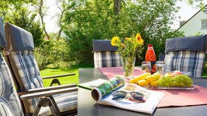 Un área de estar al aire libre con una mesa dispuesta para una comida, que incluye un libro, gafas de sol y una bandeja de fruta.