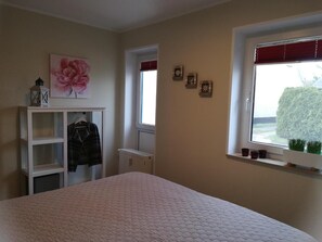 Quarto com cama, guarda-roupa, janela com vista para vegetação e parede decorada com um quadro floral e pequenos objetos.