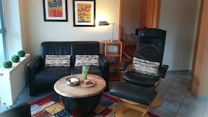 Een woonkamer met een zwarte leren bank, een zwarte leren fauteuil, een ronde houten salontafel en een kleurrijk vloerkleed. Twee ingelijste foto's hangen aan de muur, een lamp op een bijzettafeltje en een radiator met kleine planten.